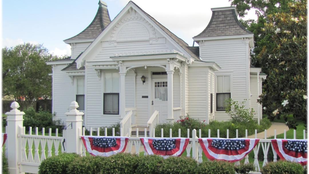 White Victorian cottage with twin turrets, ornate trim, and American flag bunting on the picket fence